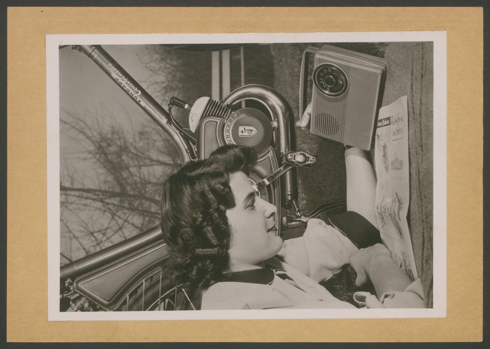 Fotografie: Nahansicht eines Zündapp-Combinette-Motors, im Vordergund lesende Frau mit Kofferradio (Stiftung Deutsches Technikmuseum Berlin CC0)