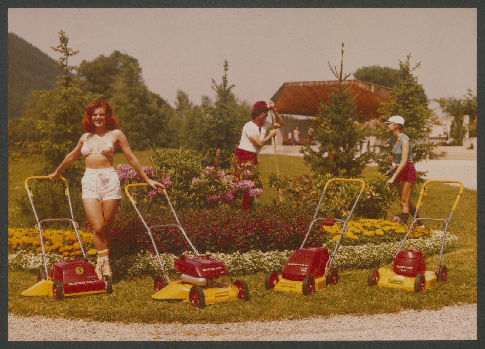 Fotografie: Vier Zündapp-Rasenmäher im Vordergrund, im Hintergrund drei Personen teilweise bei Gartenarbeit, Werbefoto (Stiftung Deutsches Technikmuseum Berlin CC0)