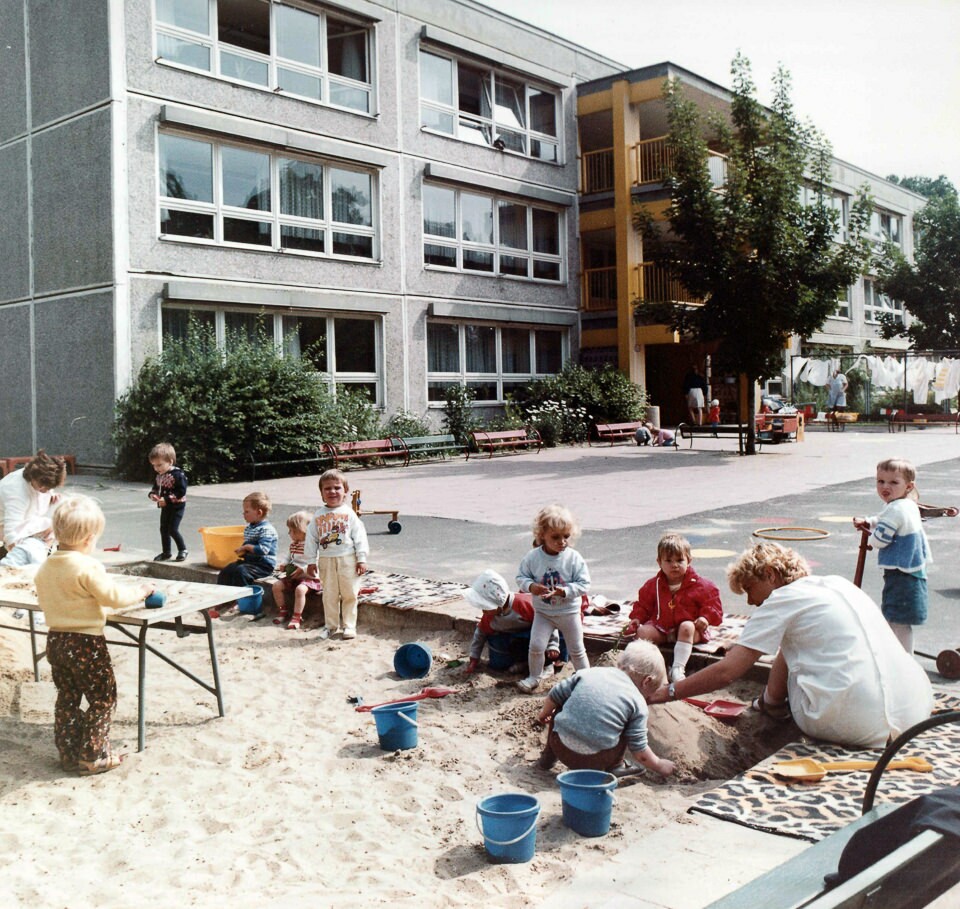 Kinder mit Erzieherin spielen im Sandkasten auf dem Spielplatz vor dem Gebäude des WF Kindergartens, Farbfoto © Kurt Schwarz, 1980er Jahre (Kurt Schwarz CC BY-NC-SA)