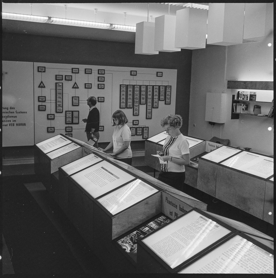 VEB Narva Kombinat, Bild 4: Drei Mitarbeiter informieren sich in einem "Ideenbank" genannten Raum des Werks mit Vitrinen und Schautafeln, zwischen den Vitr (Kurt Schwarz CC BY-NC-SA)