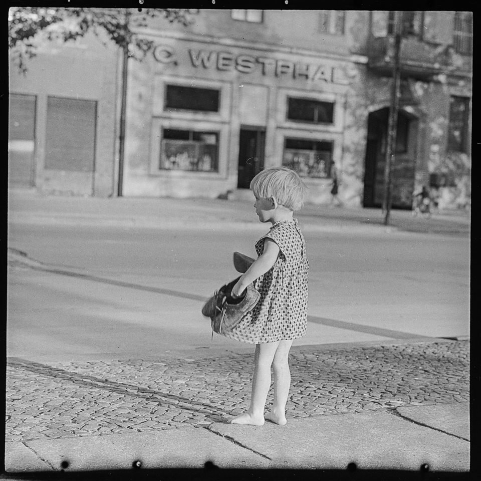Kleines Mädchen barfuß mit Schuhen in der Hand am Straßenrand. SW-Foto, 1960er Jahre © Kurt Schwarz. (Kurt Schwarz CC BY-NC-SA)