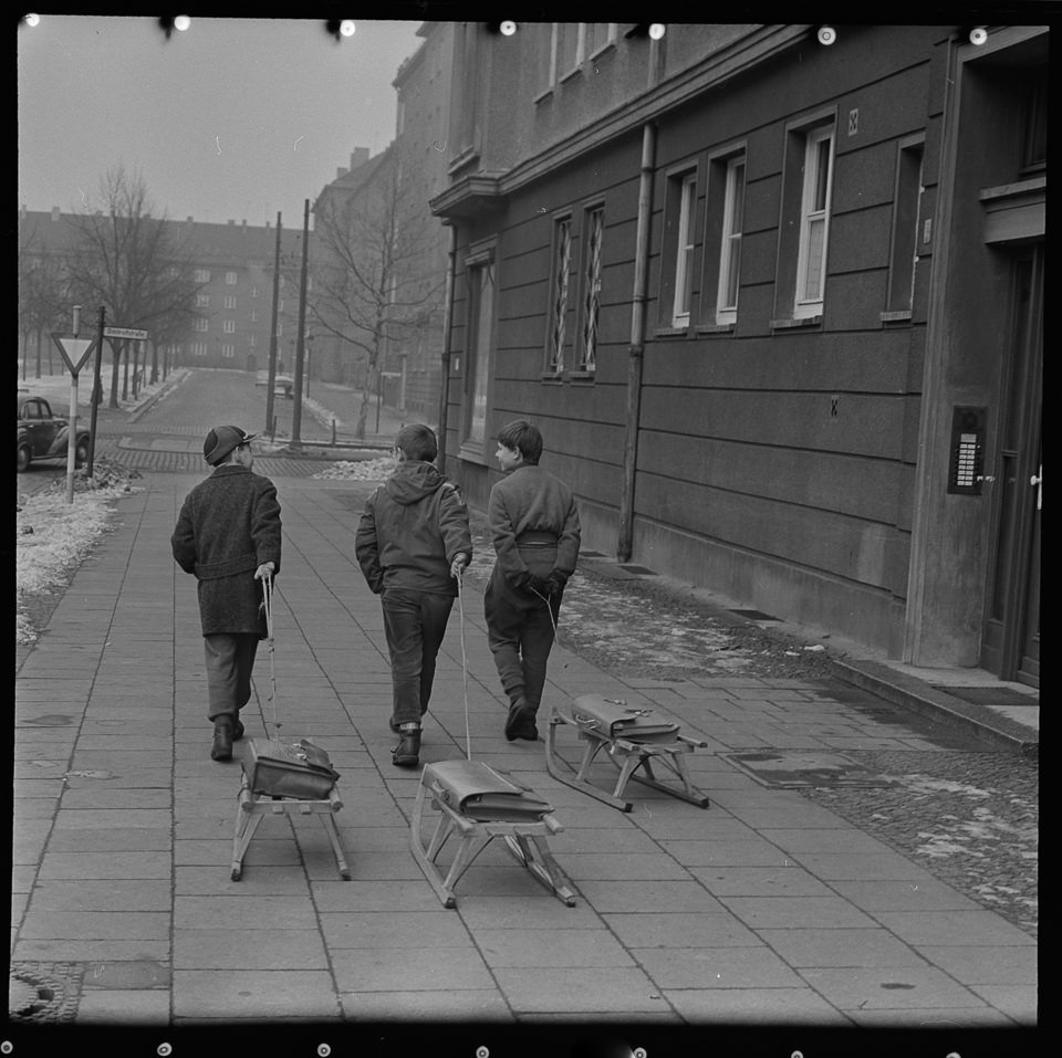 Drei Jungen ziehen Schlitten über den schneelosen Bürgersteig in einer Wohnstraße. SW-Foto, 1960er Jahre © Kurt Schwarz. (Kurt Schwarz CC BY-NC-SA)