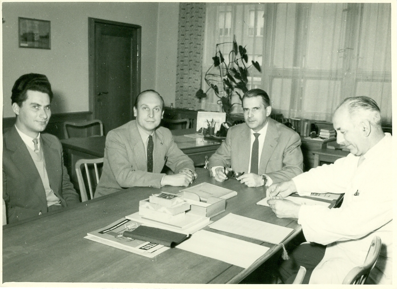 Männer in Anzügen sitzen am Schreibtisch, Tür Mitte links hinten, Fenster im Hintergrund mit Topfpflanze, Papiere auf dem Tisch (Industriesalon Schöneweide CC BY-NC-SA)