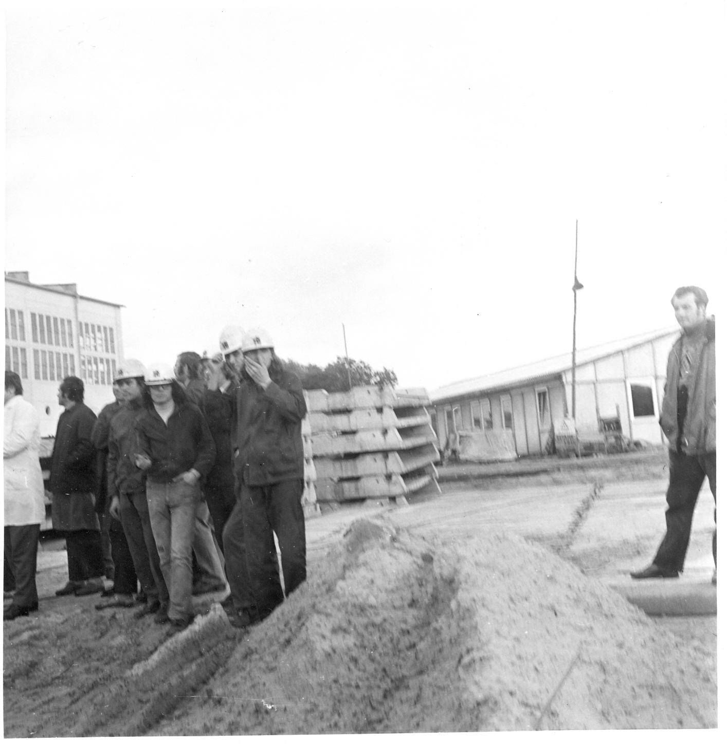 Grundsteinlegung, Arbeiter mit Helmen auf Baustelle, Sandhaufen im Vordergrund, im Hintergrund Barracke und Betonteile (Industriesalon Schöneweide CC BY-SA)