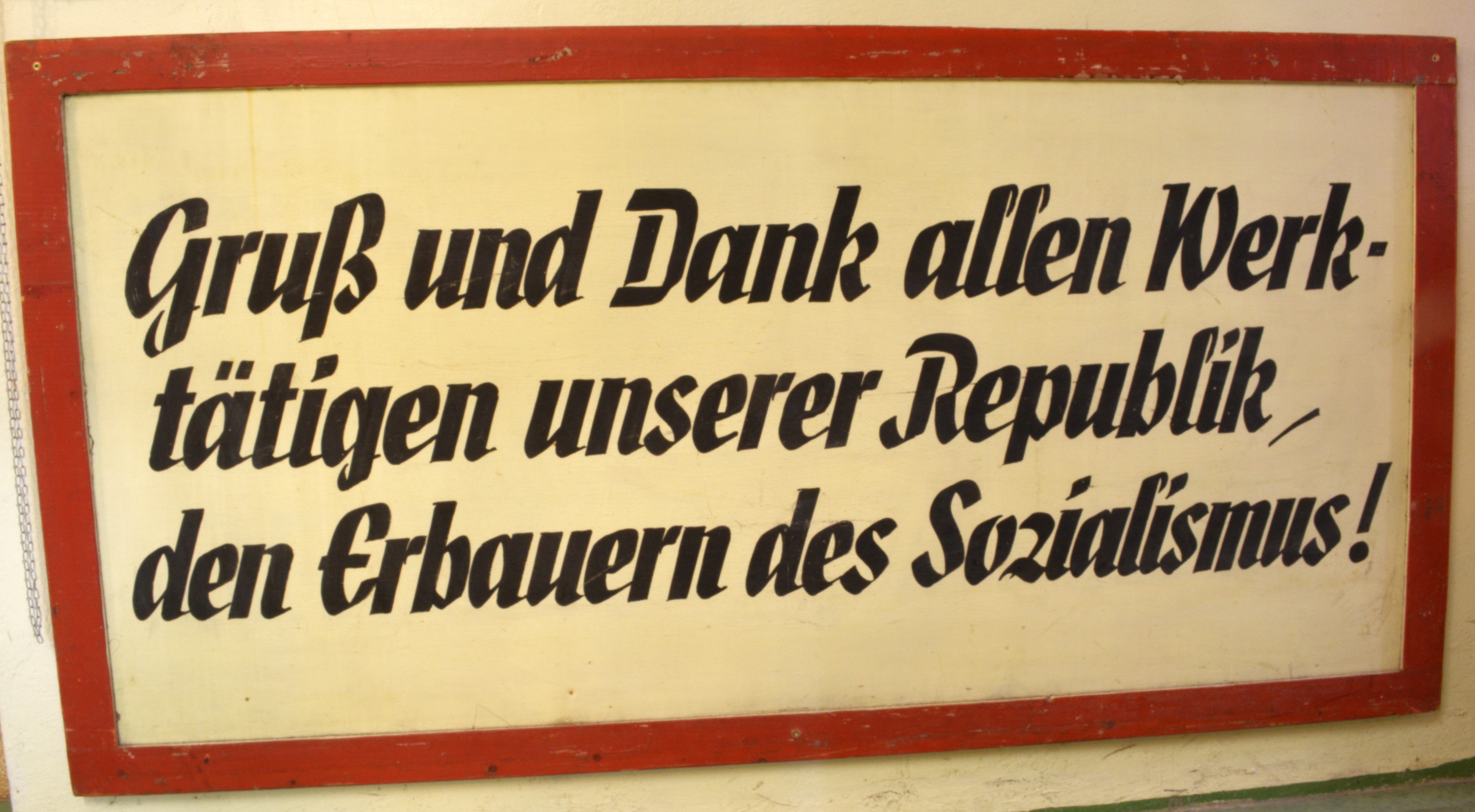 Schild mit Parole: "Gruß und Dank allen Werktätigen unserer Republik, den Erbauern des Sozialismus!" (DDR Geschichtsmuseum im Dokumentationszentrum Perleberg CC BY-SA)