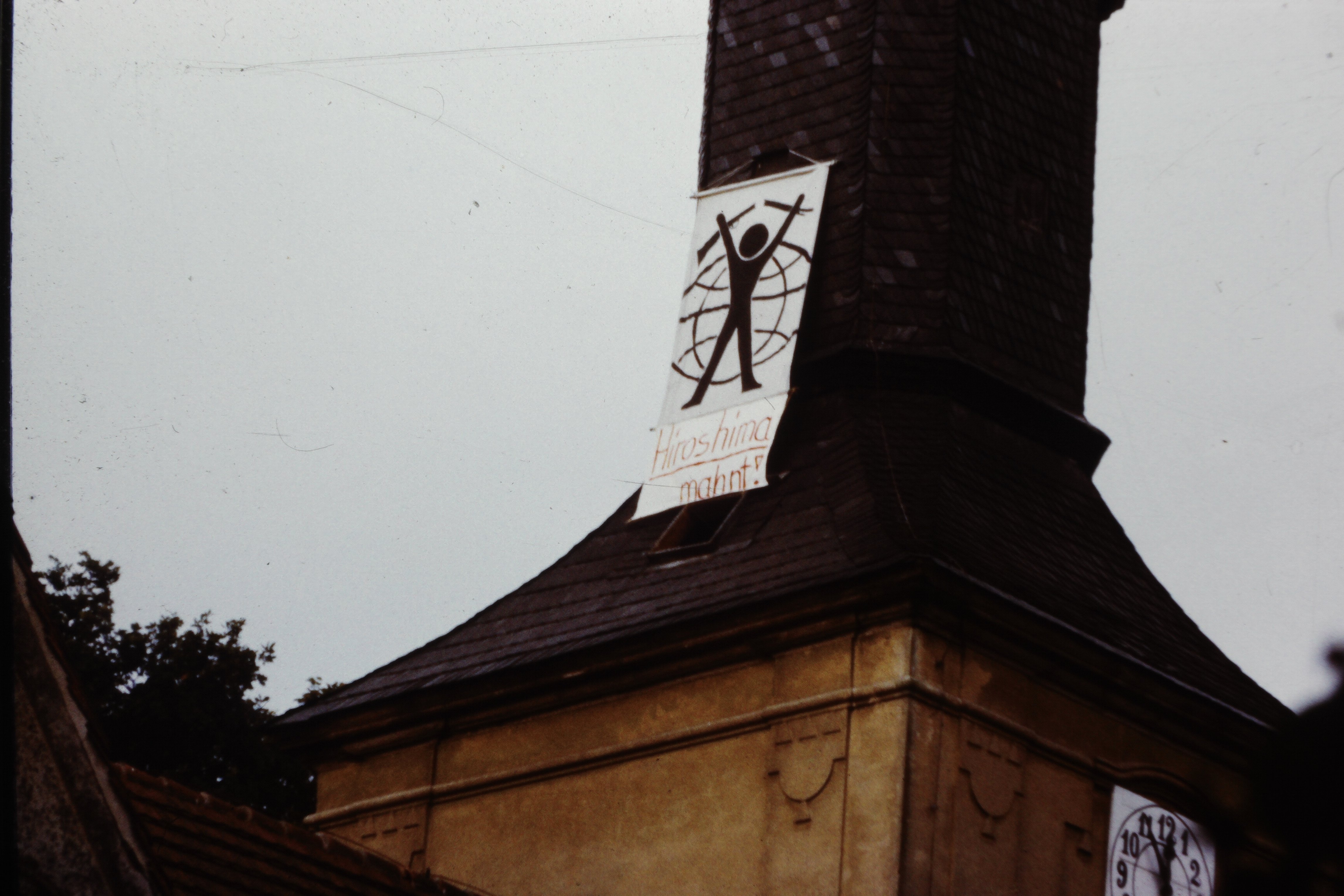 Kirchturm der Köritzer Gemeinde in Neustadt (Dosse): Transparent mit aufgemaltem "Mann vor Weltkugel" und der Aufschrift "Hiroshima mahnt!" (DDR Geschichtsmuseum im Dokumentationszentrum Perleberg CC BY-SA)