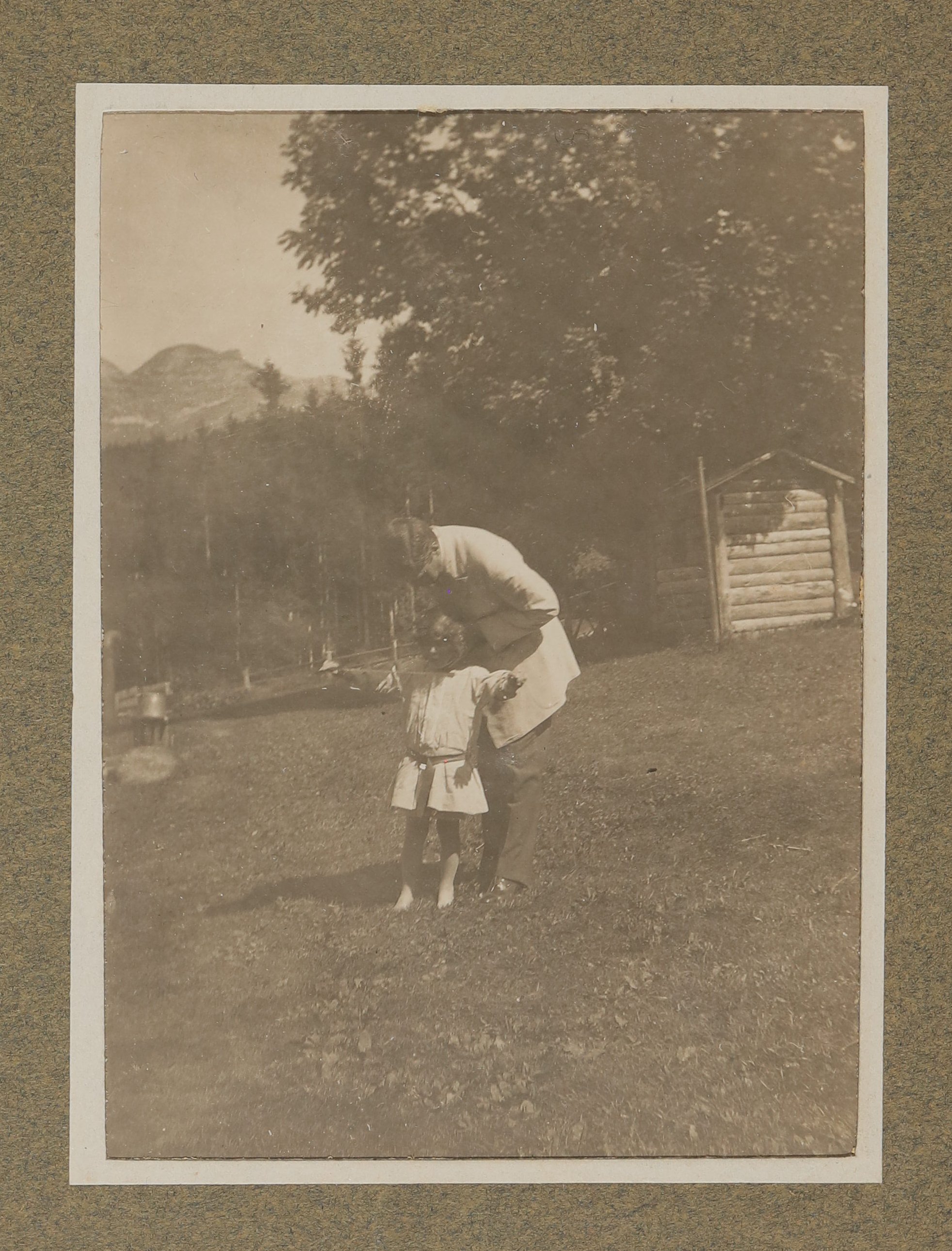 Hugo von Hofmannsthal mit einem Kind (Raimund oder Christiane?) im Garten vor Bergen stehend, sich zum Kind hinunterbeugend (Vera Graaf CC BY-NC-SA)