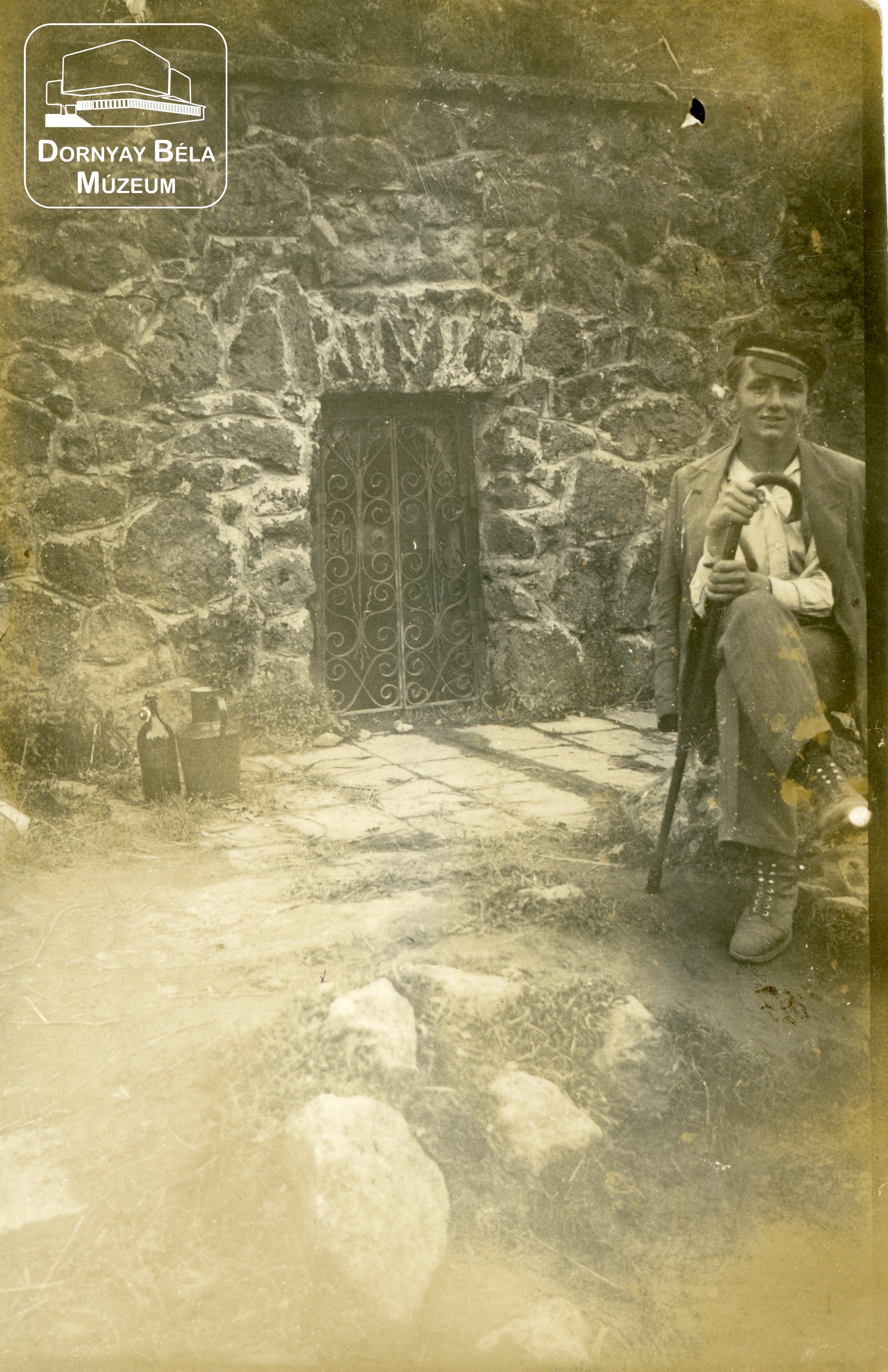 Dr. Dornyay Béla gyűjteményéből: Kövön ülő fiatal férfi egy vasrácsos bejárat előtt. (Dornyay Béla Múzeum, Salgótarján CC BY-NC-SA)
