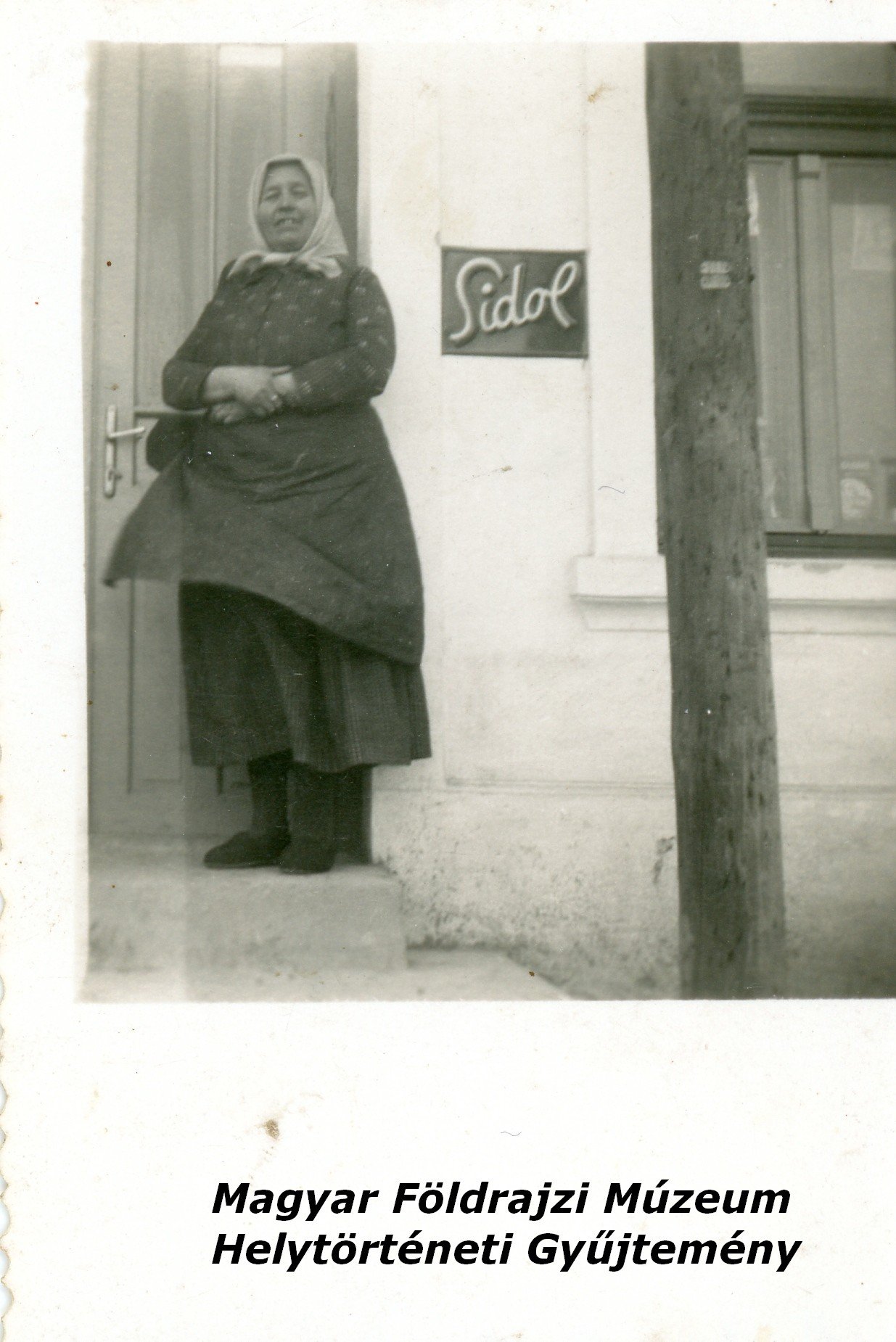 Fejkendős asszony a Gyurcsek vegyesbolt ajtajában. (Magyar Földrajzi Múzeum CC BY-NC-SA)