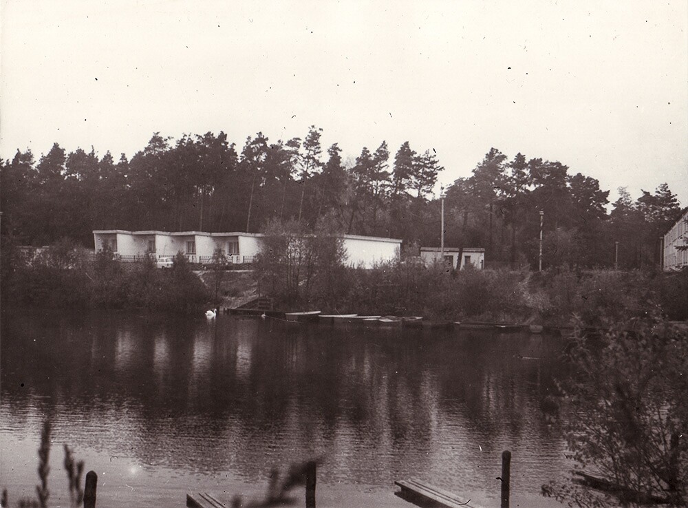 Bungalows im Wald am See, im Vordergrund Boote und ein Schwan auf dem Wasser (Museum Schloss Bernburg / Deutsche Stiftung Denkmalschutz CC BY-NC-SA)